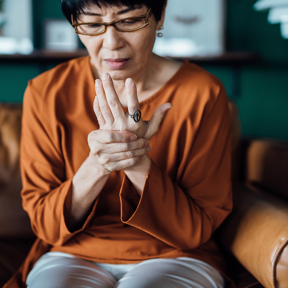 Senior Asian woman with joint pain in her hand while sitting on sofa at home