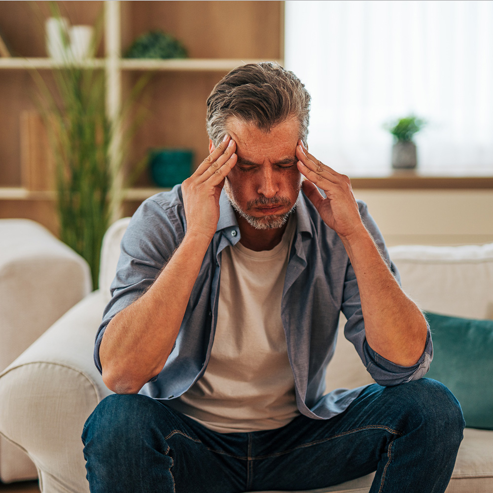 Man sitting on the sofa in living room, holding his head in his hands, grappling with headache pain from posture